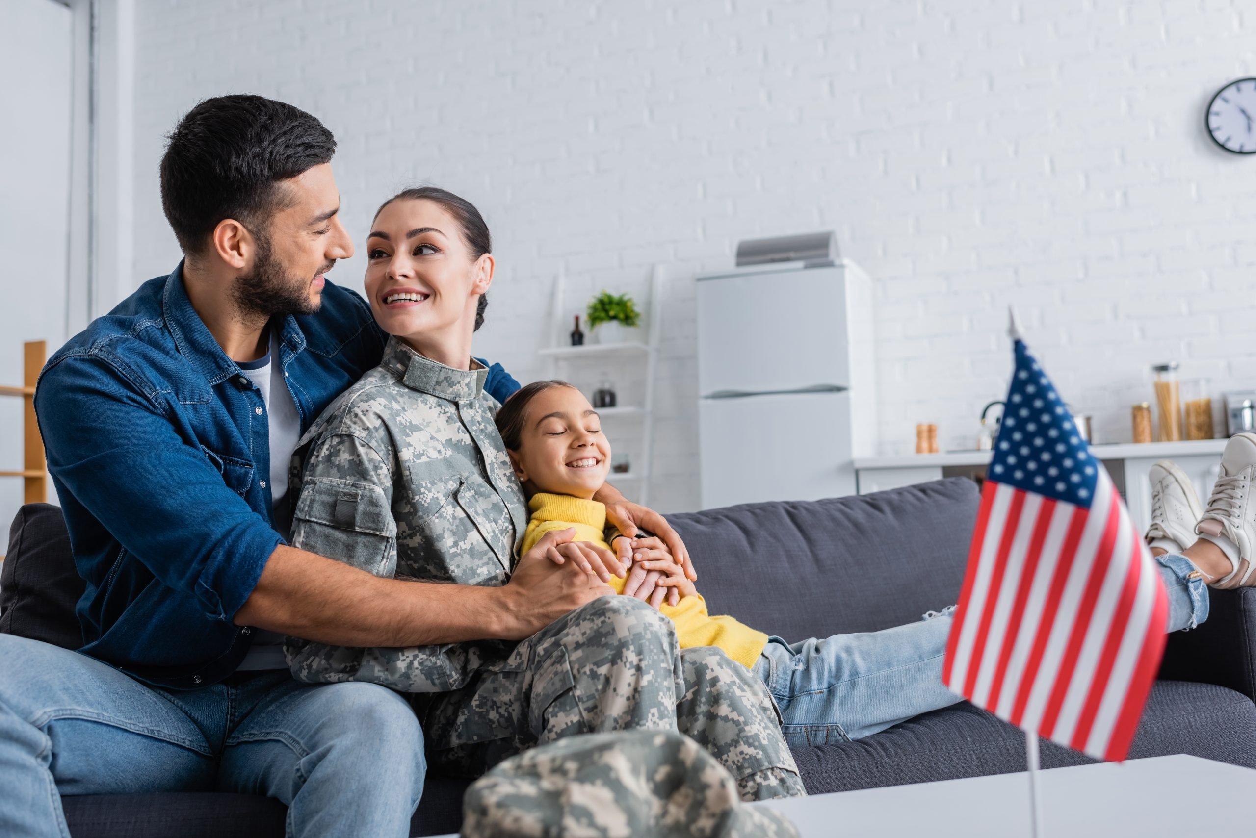 Positive man hugging wife in camouflage uniform near daughter and blurred american flag at home